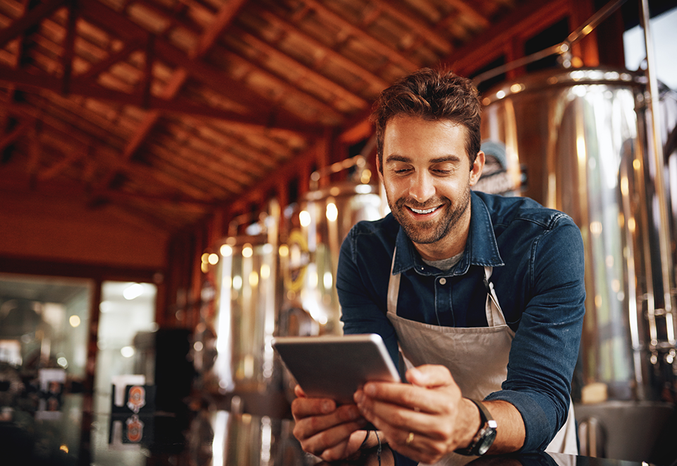 Sm Bus Loans - 960x660 – Chemung Canal Trust Company  A man in an apron leans on the counter, reviewing a tablet