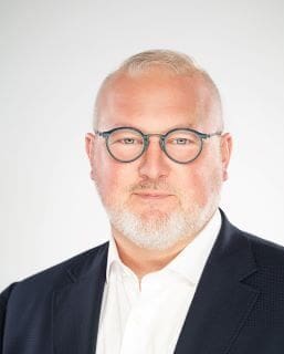 Portrait of a middle-aged man with gray hair and beard wearing rectangular glasses, a white shirt, and a dark blazer, looking at the camera against a light background.
