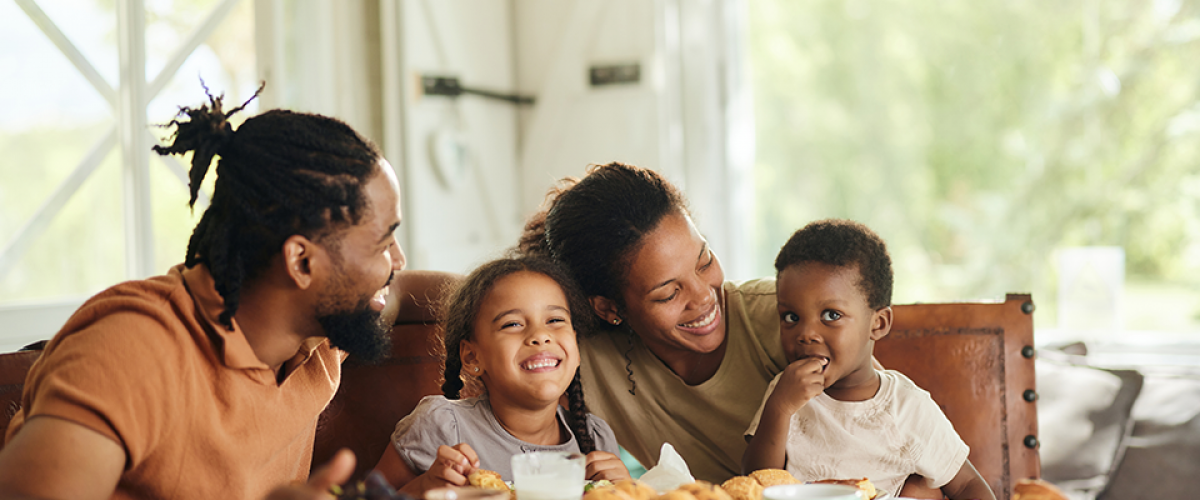 A family of four - two parents and two young children, sit at their table with breakfast in front of them