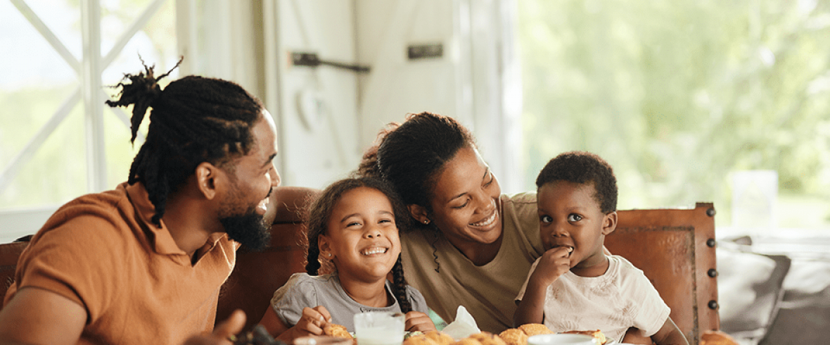 A family of four - two parents and two young children, sit at their table with breakfast in front of them