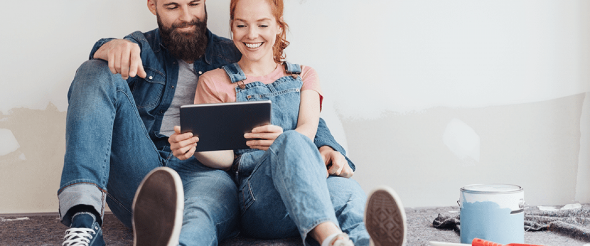 A couple is seated in front of their unfinished wall, holding a tablet. There are cans of paint and supplies next to them